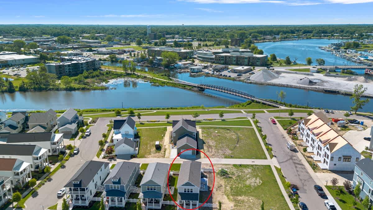 Aerial view of a modern residential development with multiple houses, a winding river, marina, and distant city buildings under a clear blue sky. A red circle highlights a specific property within the neighborhood.