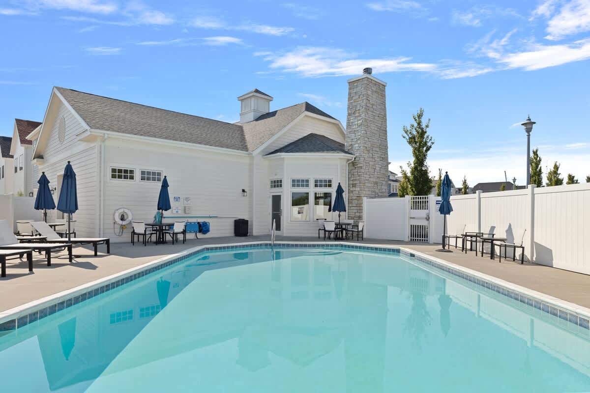 A sparkling rectangular outdoor swimming pool surrounded by a concrete deck with lounge chairs and dining tables with umbrellas, set against a backdrop of a clubhouse building.