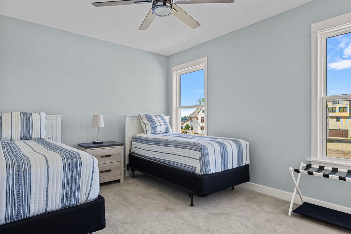 A bright bedroom with light blue walls, featuring two twin beds with striped blue and white bedding, a white nightstand with a lamp, and a large window offering a view of other houses.
