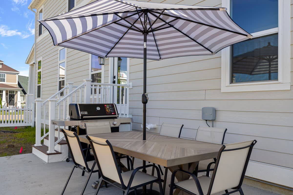 An outdoor patio area with a large rectangular dining table and six chairs under a striped umbrella, adjacent to a Weber grill and the light-colored siding of the house.