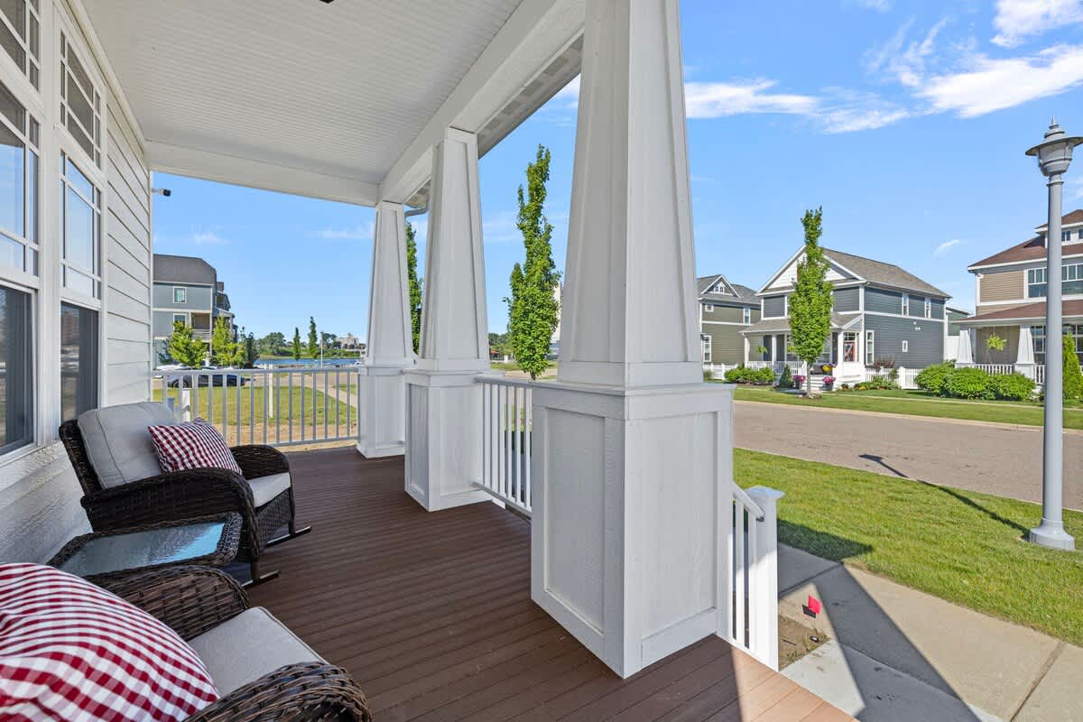 A cozy front porch with dark wood flooring, white columns, and railings, furnished with two wicker rocking chairs with red and white checkered pillows, offering a view of the neighborhood street.