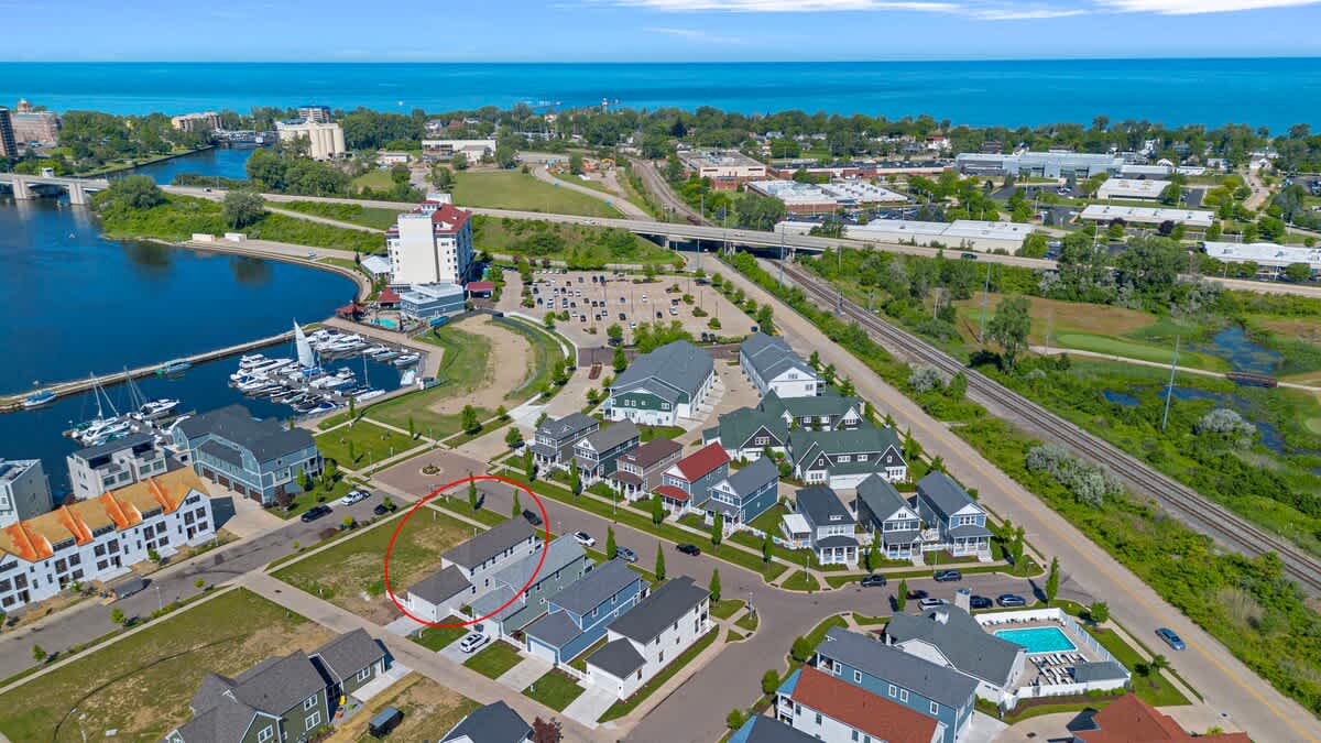 Expansive aerial view showcasing a modern residential community adjacent to a river and marina, with a large body of water (likely a lake) and distant shoreline visible under a bright blue sky. A red circle highlights a specific house.