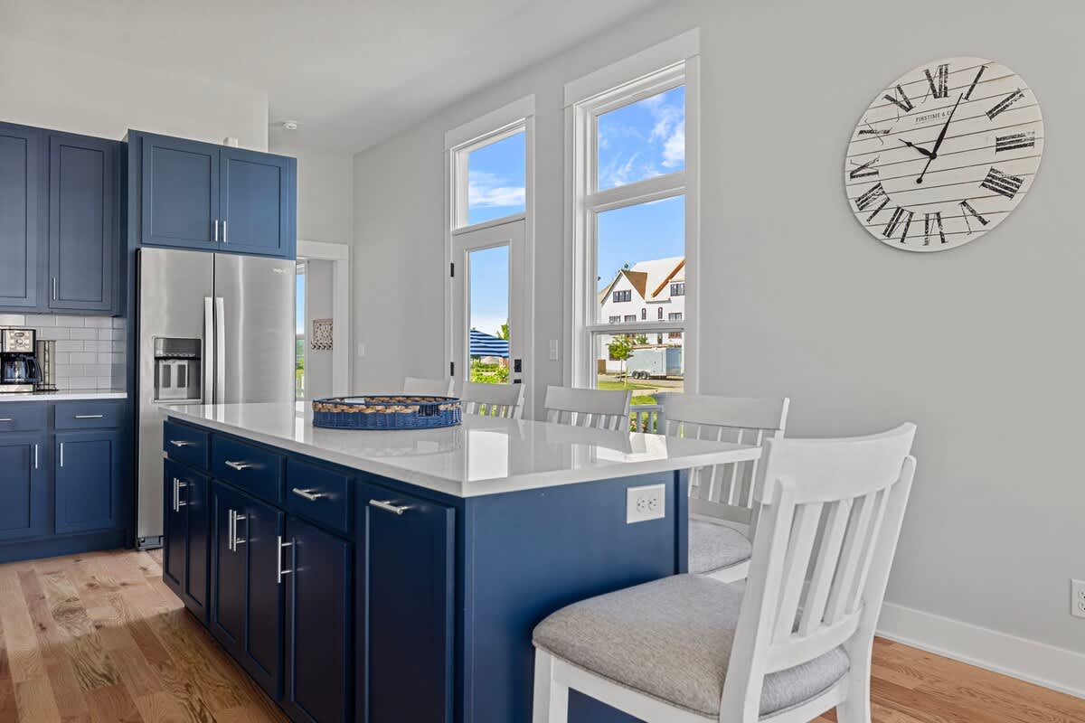 Modern kitchen featuring a large dark blue island with a white quartz countertop and four white bar stools, surrounded by light gray walls and tall windows, providing a bright dining space.
