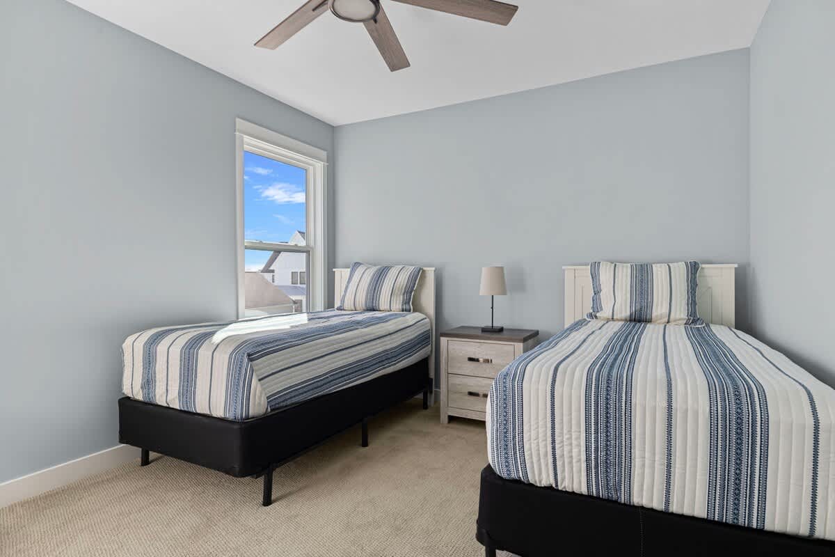 A serene bedroom with light blue walls, showcasing two twin beds with matching blue and white striped bedding, a white nightstand with a lamp, and a window providing natural light.