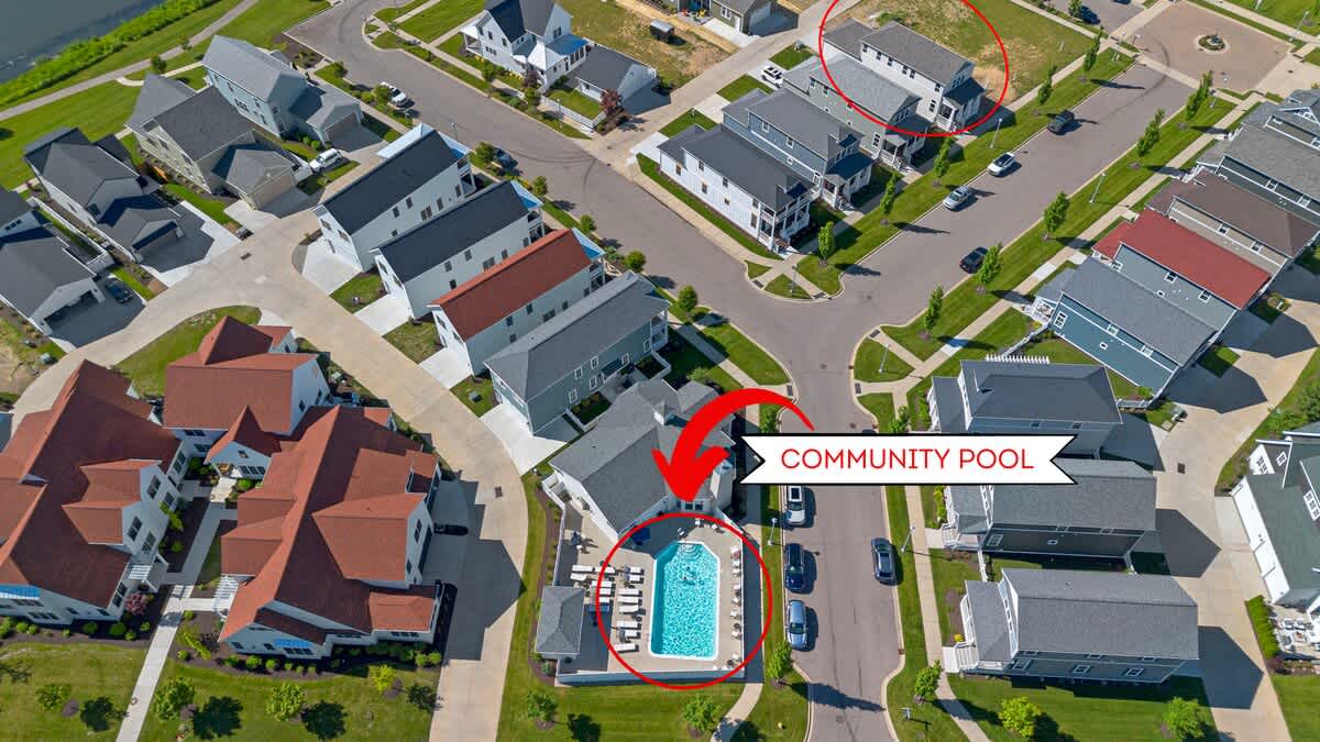 Overhead aerial view of a modern residential community featuring a rectangular community pool with lounge chairs, surrounded by contemporary houses with varying rooflines. A red arrow points to the pool, and a red circle highlights another house.
