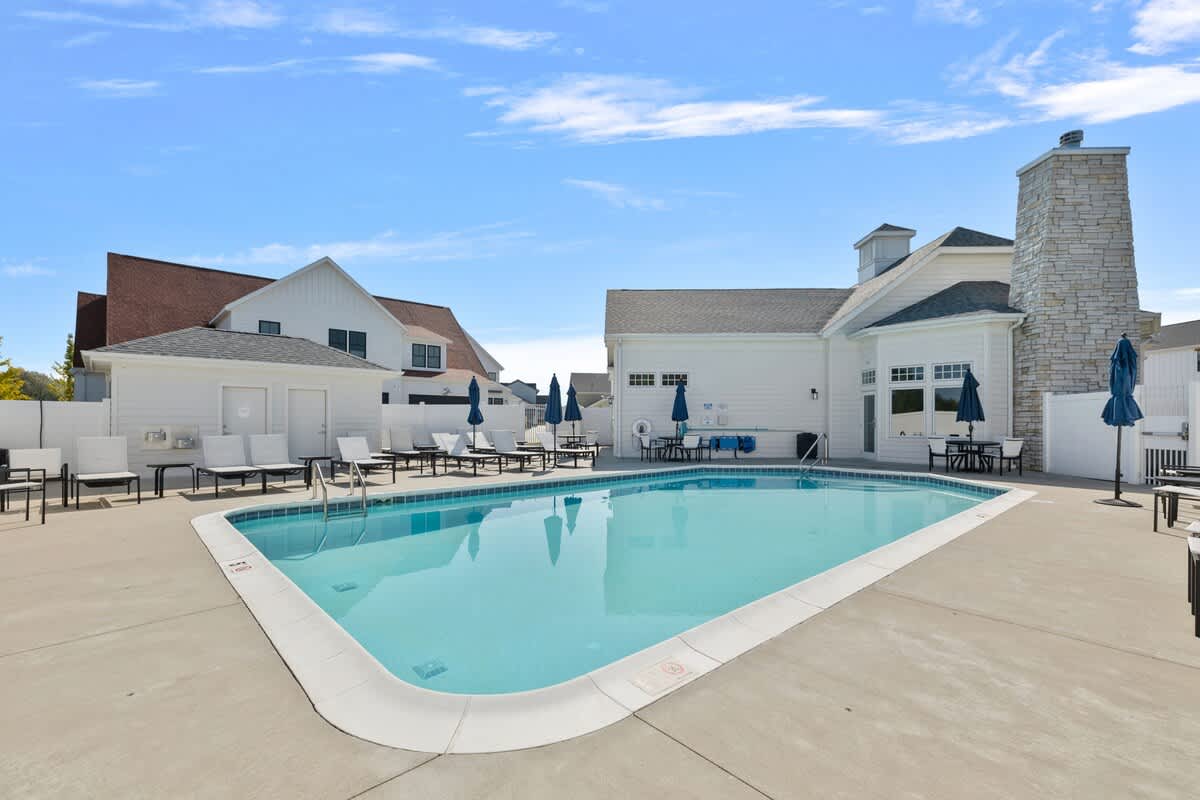A wide view of the community outdoor swimming pool, featuring a large concrete deck with multiple lounge chairs and shaded dining areas, with the clubhouse building and a stone chimney in the background.