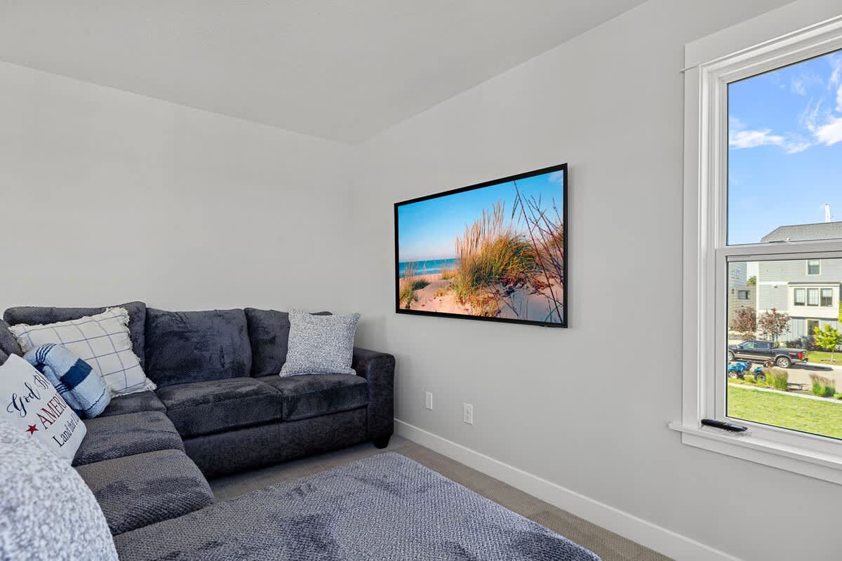 Cozy media room featuring a large dark gray sectional sofa with decorative pillows, a wall-mounted TV displaying a beach scene, and a large window.