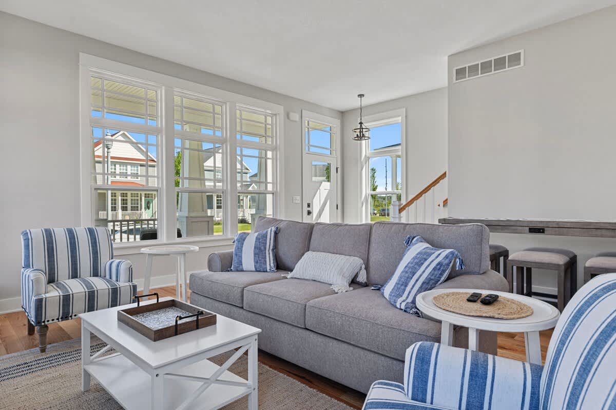 Cozy living room featuring a gray sofa with blue and white striped pillows, two matching armchairs, a white coffee table, and large windows providing ample natural light and views of the neighborhood.