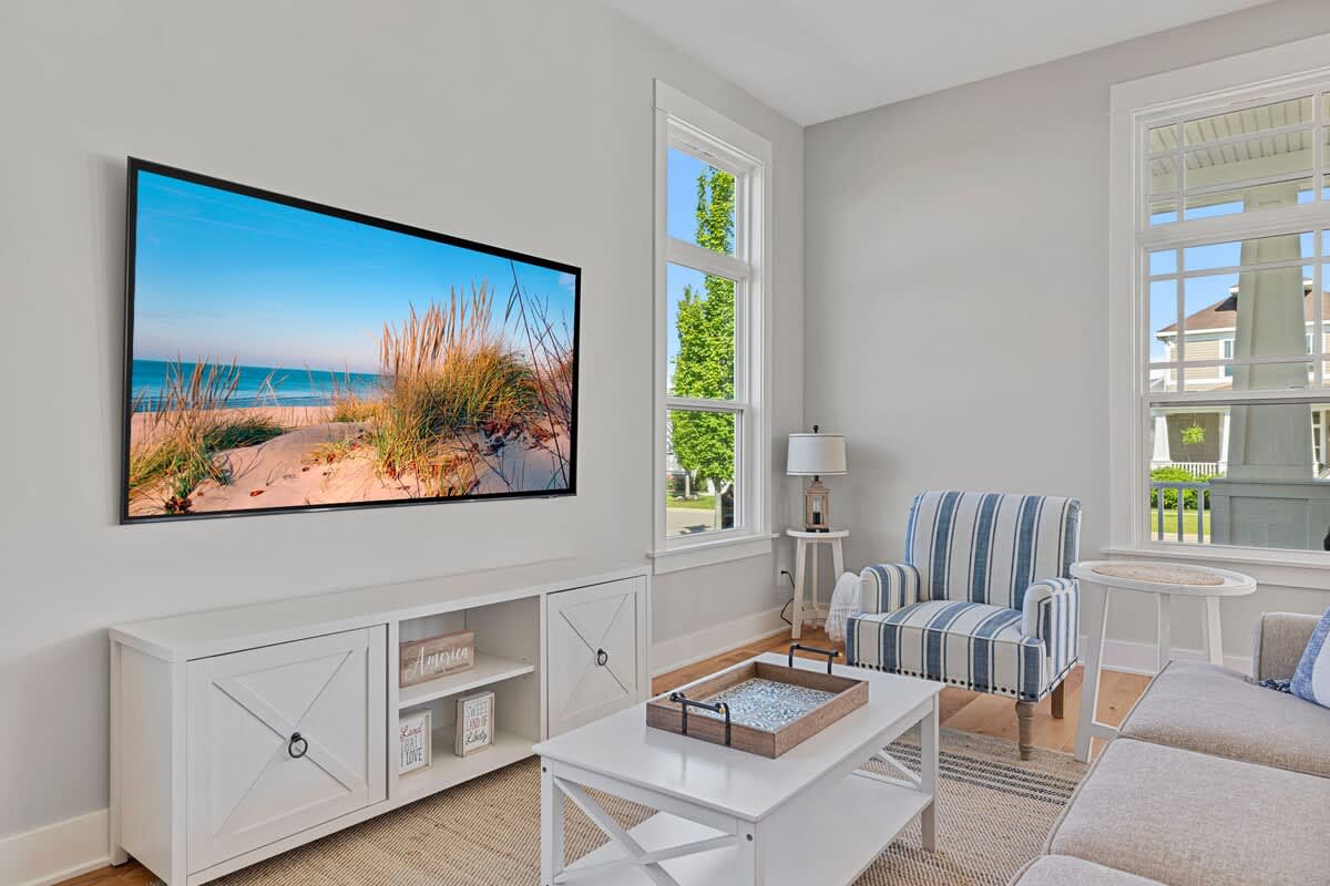 Bright living room with a large wall-mounted TV above a white media console, a striped armchair, and tall windows, creating a comfortable space for relaxation.
