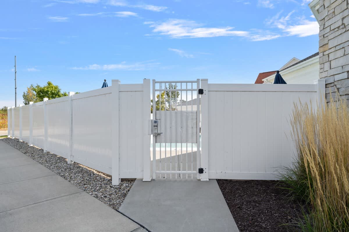 A white privacy fence with a coded gate provides access to a community pool area, with glimpses of the pool and surrounding buildings beyond the fence.