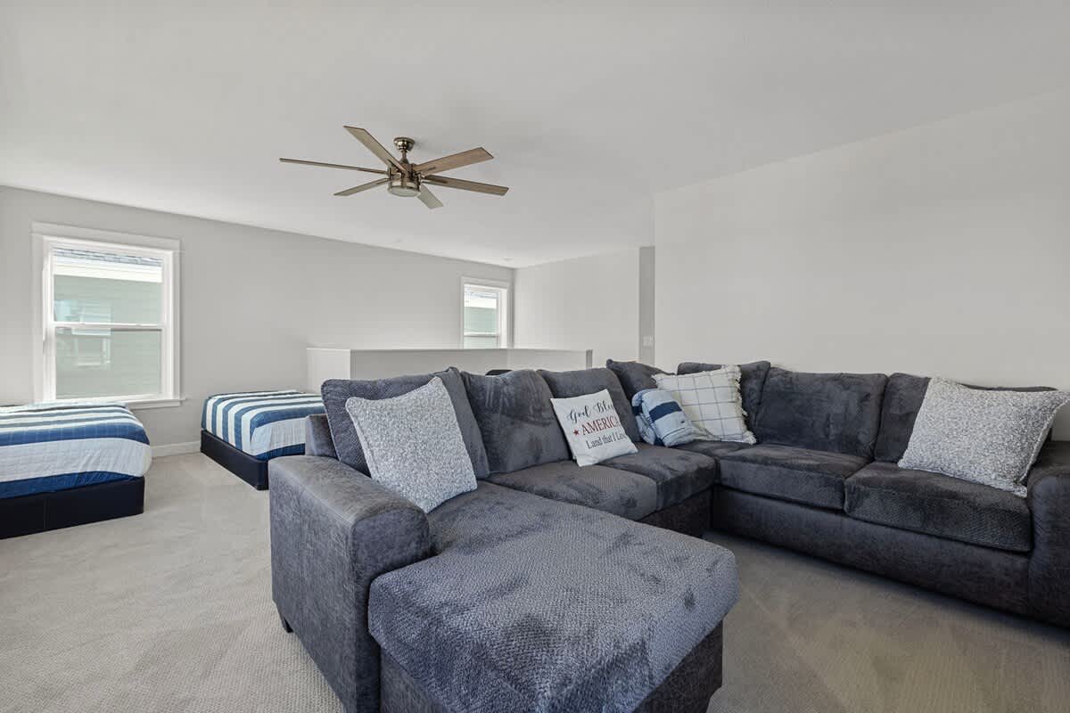 Spacious media room or secondary living area with a large dark gray sectional sofa, a ceiling fan, and two twin beds with striped bedding visible in the background.