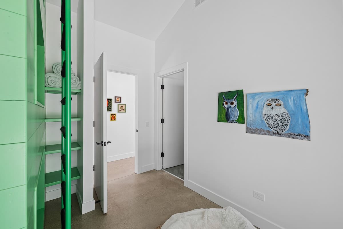 A bright hallway with white walls and concrete flooring, featuring colorful owl-themed artwork on the wall. An open doorway leads to another room, and the green bunk bed structure is partially visible on the left.