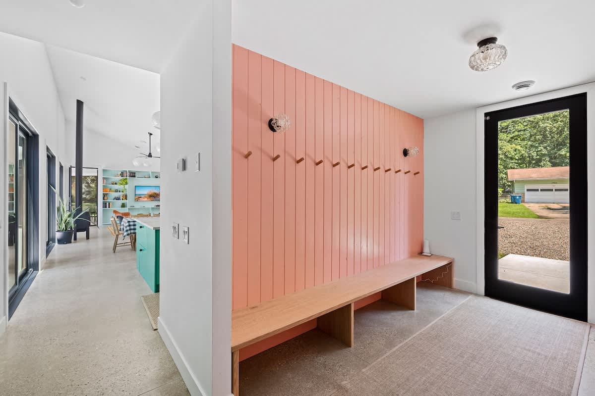 A welcoming entry area featuring a vibrant peach-colored slatted accent wall with numerous coat hooks and a long wooden bench. The space has a modern aesthetic with a black-framed glass door leading to the exterior.