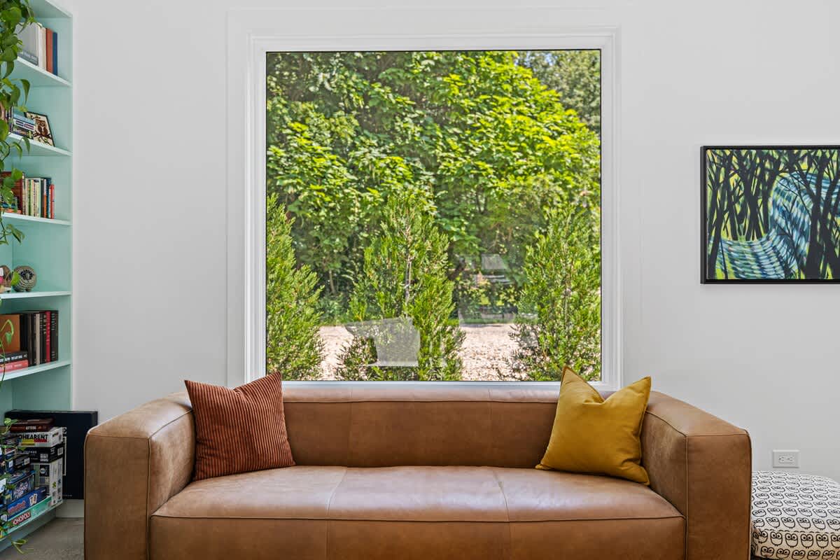 A comfortable brown leather sofa with accent pillows is positioned in front of a large window, offering a serene view of lush green trees and shrubs outside. Part of a teal built-in bookshelf is visible on the left.