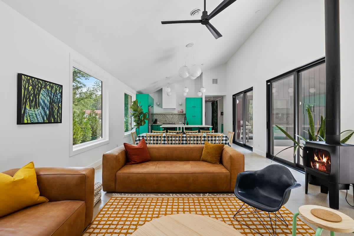 A spacious, bright living room featuring a tan leather sofa, a black wood-burning stove, and a large area rug with a yellow grid pattern. The open-concept space extends to a kitchen with vibrant teal cabinetry and a dining area.