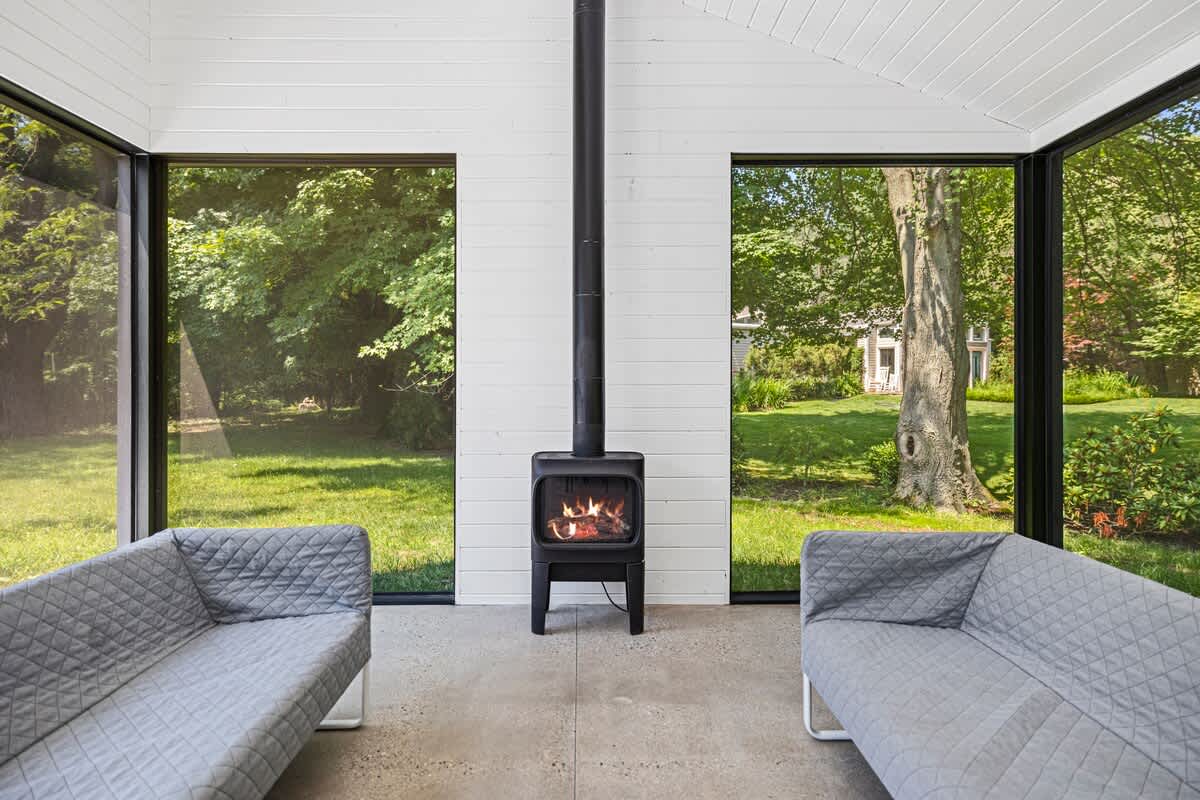Cozy sunroom with white paneled walls and a vaulted ceiling, featuring a modern black wood-burning stove centered between two large windows offering views of a lush green backyard. Two grey quilted sofas face each other.