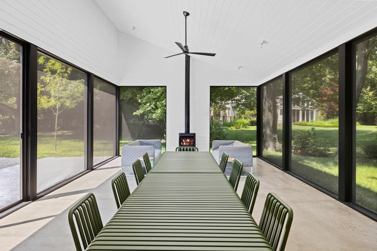 Contemporary dining space featuring a long green slatted table and chairs, concrete flooring, and expansive windows overlooking lush greenery. A sleek black wood-burning stove stands centrally.