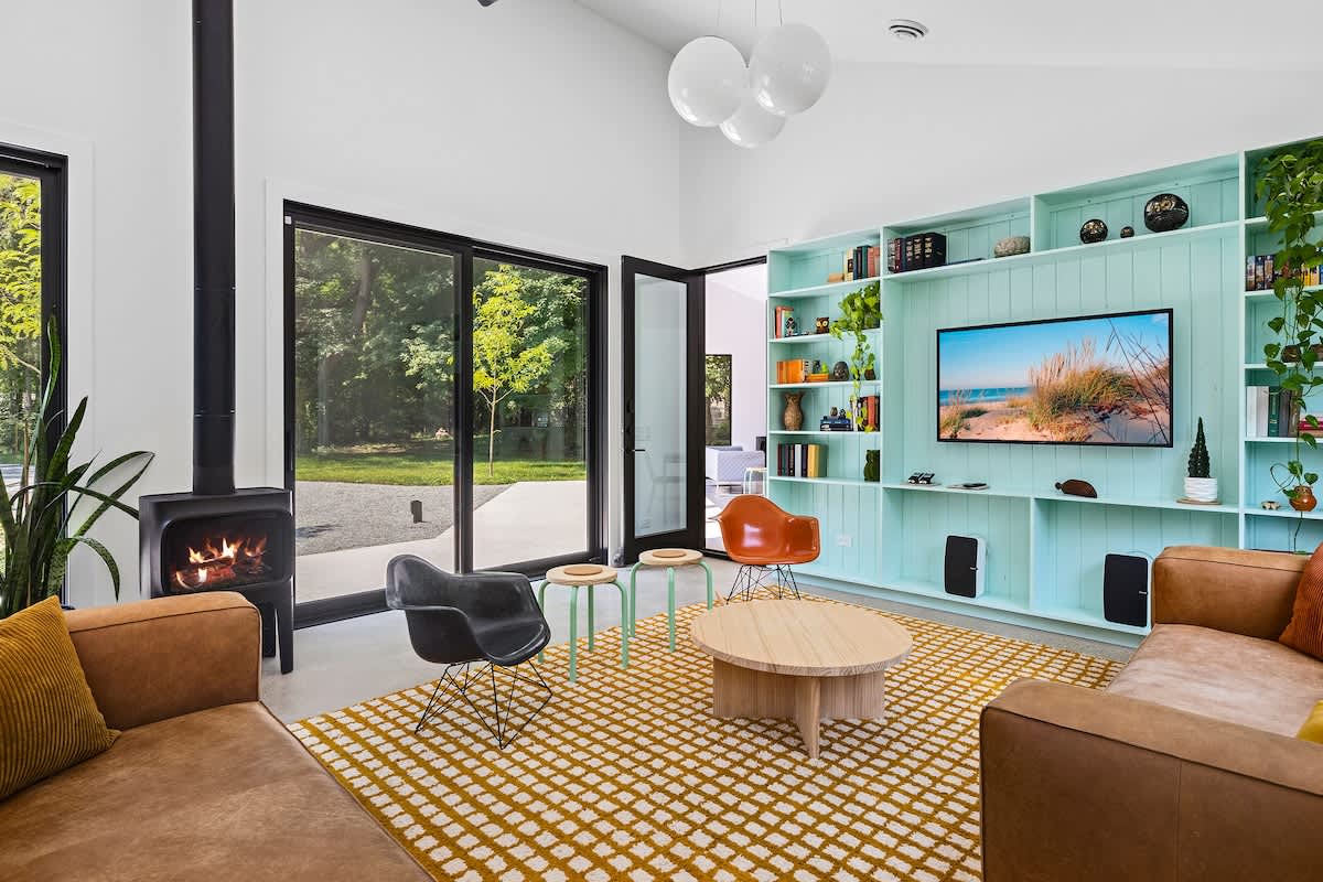 A bright and spacious living room with a black wood-burning stove, two brown leather sofas, and a large yellow patterned rug. A teal built-in entertainment unit with a TV and decor lines one wall, and sliding glass doors open to the exterior.