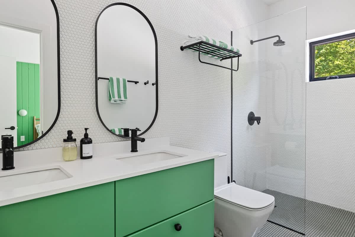 Modern bathroom with a double vanity featuring bright green cabinets and white countertops, complemented by black oval mirrors and fixtures. The walls are covered in white penny tiles, and a walk-in shower with a glass enclosure is visible.