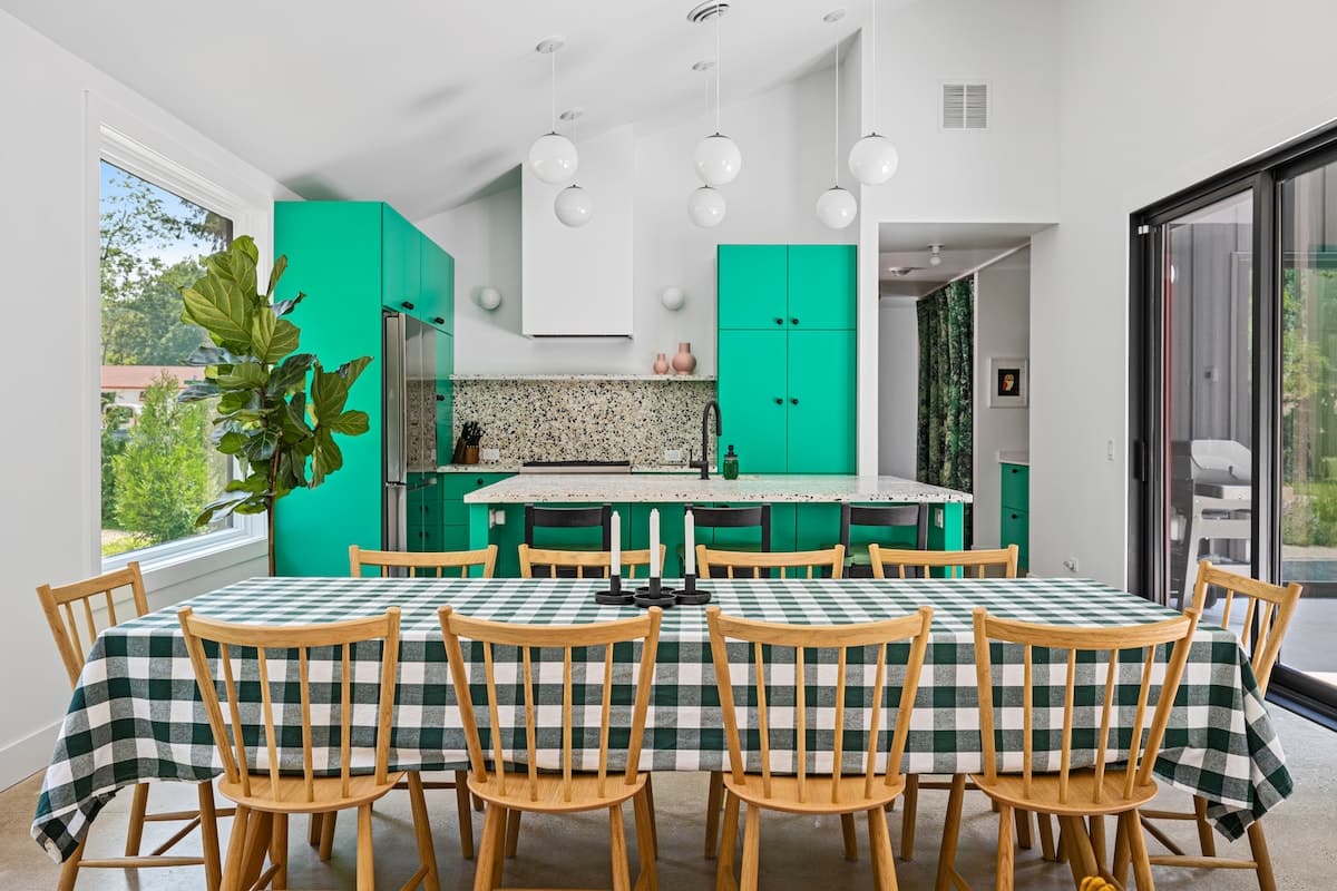 Spacious dining area featuring a long wooden table with a green and white checkered tablecloth, surrounded by wooden chairs, adjacent to a modern kitchen with bright green cabinetry and white globe pendant lights.
