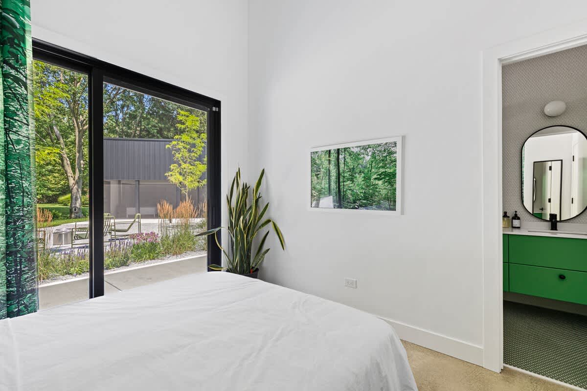 Bright bedroom with a white bed, large sliding glass doors opening to a patio and garden, and a vibrant green forest-print curtain. A snake plant adds a touch of greenery, and a modern bathroom is visible through an open doorway.