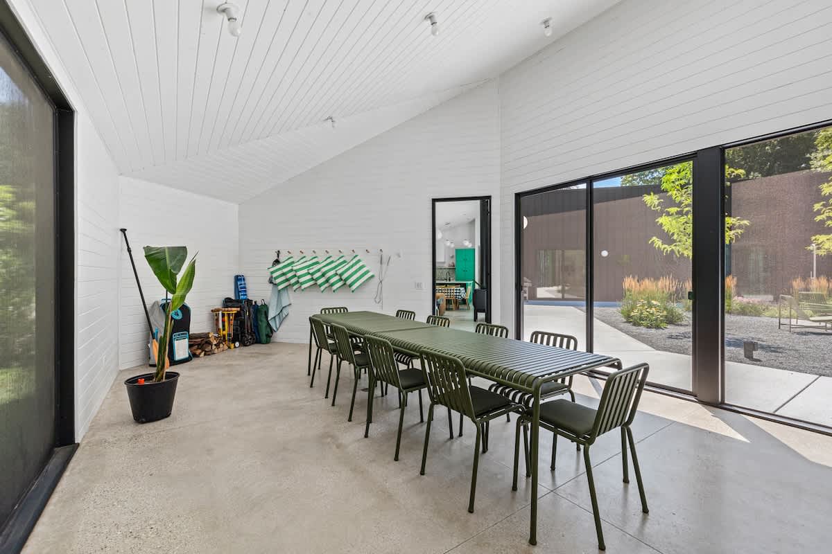 Spacious dining room with a long green slatted dining table and matching chairs, set against white shiplap walls and a concrete floor. Large sliding glass doors open to an outdoor area, and a row of green towels hangs on the wall.