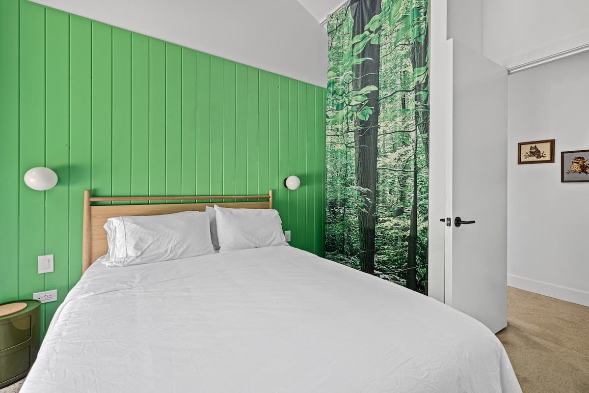 Another perspective of the bedroom, highlighting the green shiplap accent wall and the simple wooden bed with white linens. A small green nightstand with a drawer is visible, along with modern wall sconces.