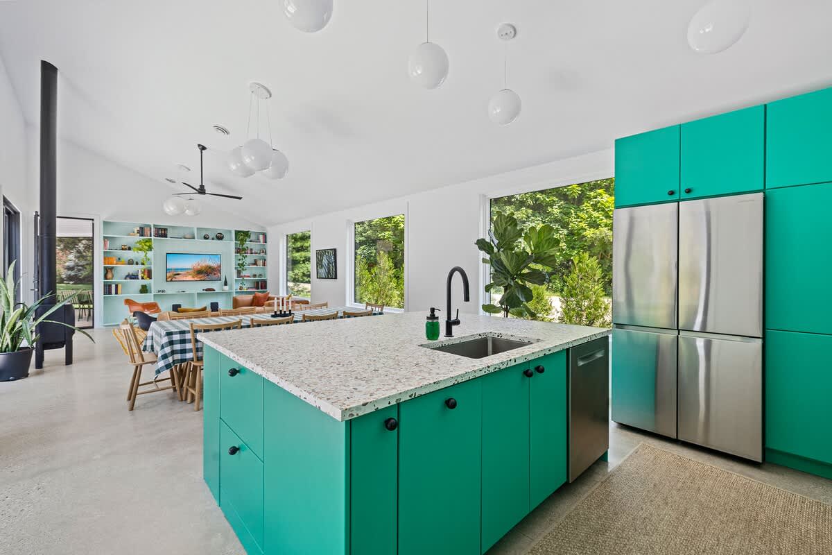Spacious open-concept kitchen featuring bright green cabinets, a large stainless steel refrigerator, and a terrazzo island. The space flows into a living area with built-in shelving.