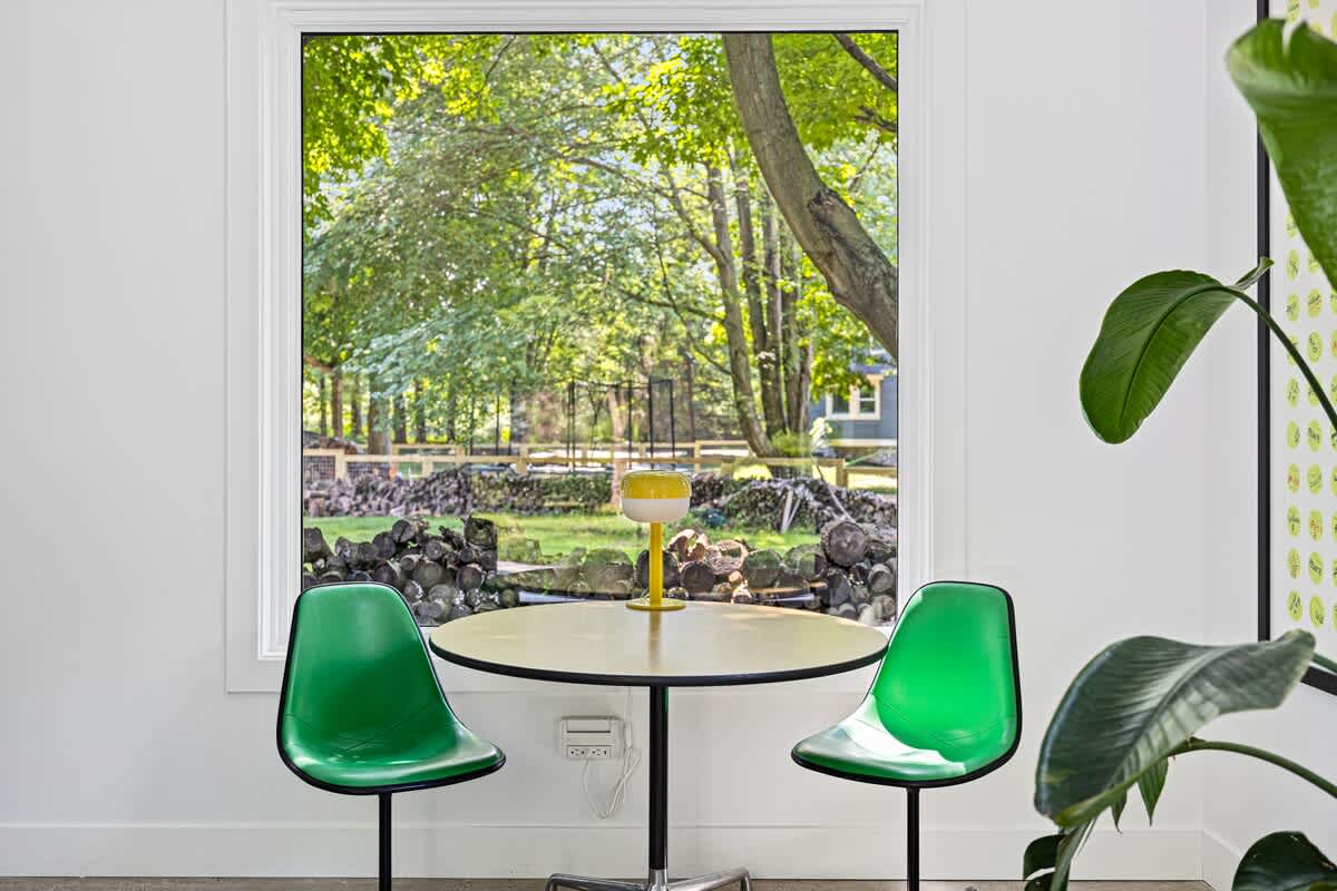 Bright breakfast nook with a small round table and two vibrant green chairs, positioned by a large window offering a view of a wooded area with stacked logs. A yellow lamp sits on the table.