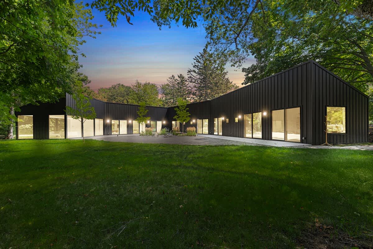 Expansive exterior view of the modern black-clad house, showcasing its unique curved architecture and numerous large windows and sliding doors opening to a central grassy courtyard. Mature trees frame the property under a twilight sky.