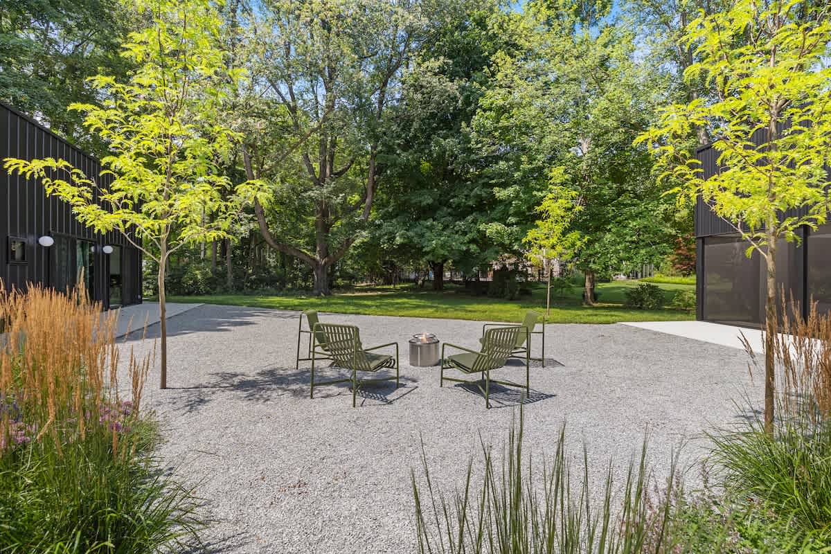 Outdoor gravel patio area surrounded by lush greenery and trees, featuring four modern green chairs arranged around a central fire pit. The property's black exterior is visible in the background.