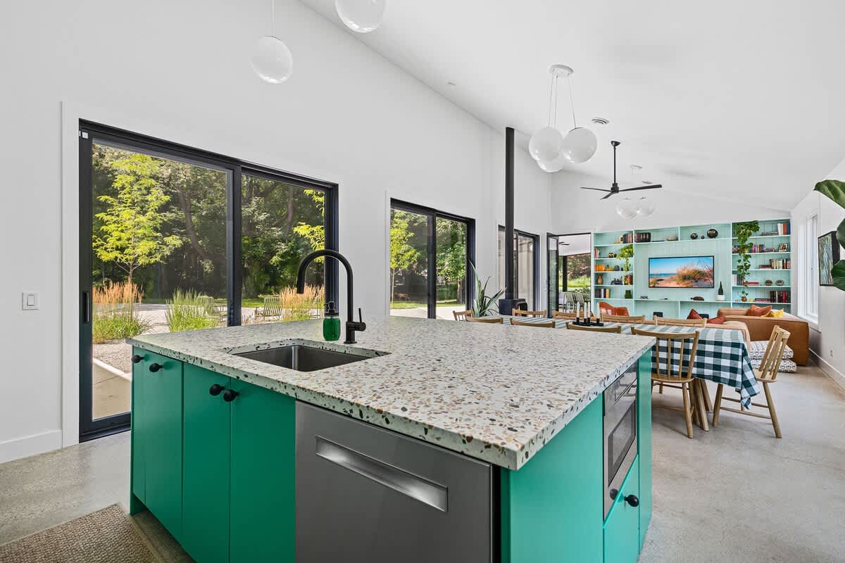 Modern kitchen island with a built-in sink, black faucet, and a speckled terrazzo countertop, featuring bright green cabinetry below. Large windows offer views of the outdoors.