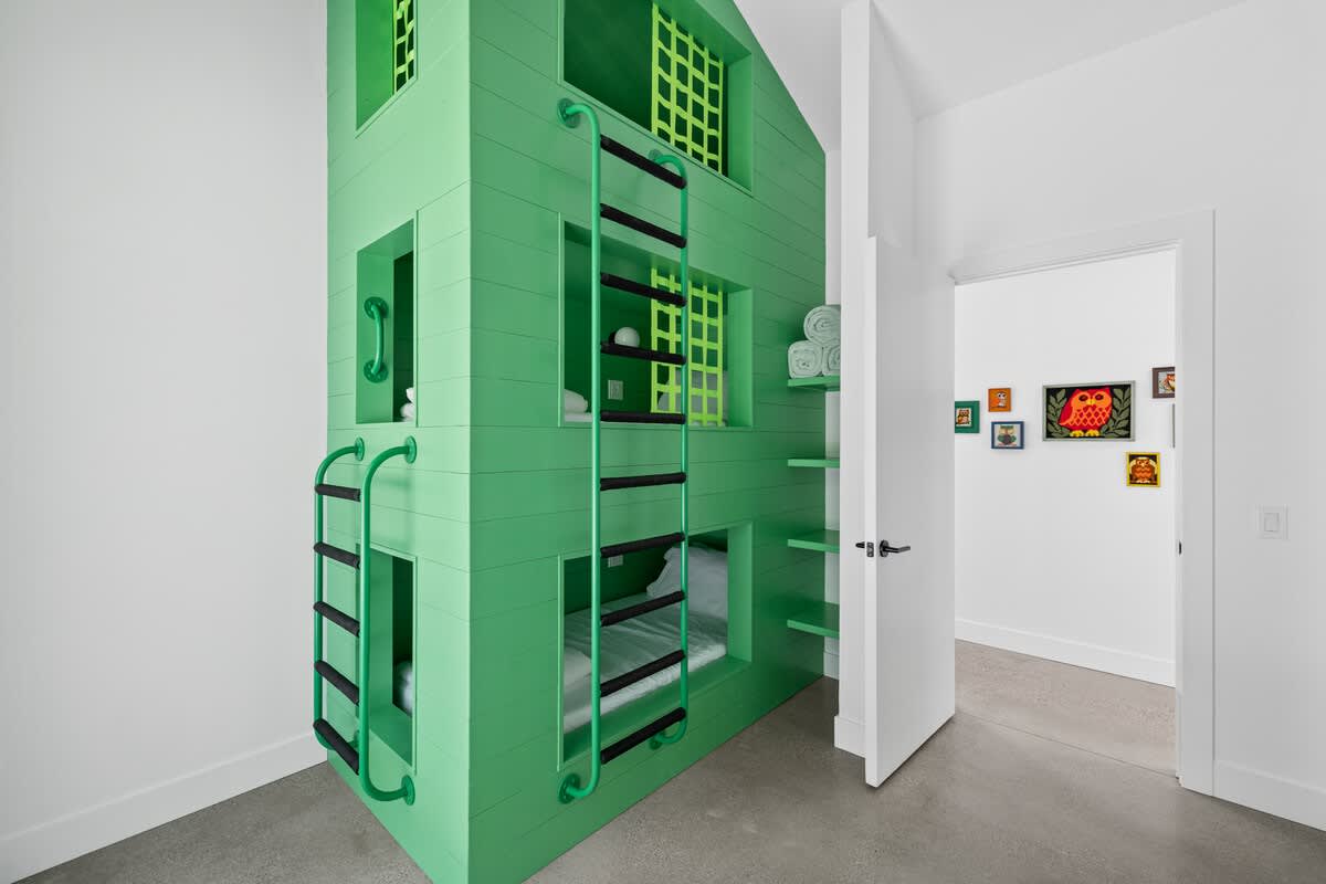 Another view of the vibrant green custom bunk bed structure, showcasing its multi-level design with black ladders and integrated shelving. The room has white walls and concrete flooring, with an open doorway leading to a hallway.