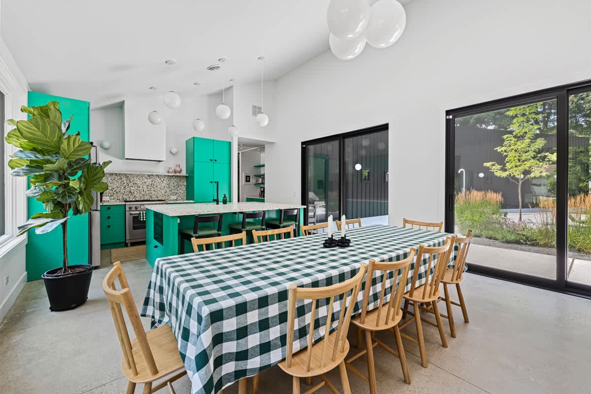 Bright dining area with a green and white checkered tablecloth on a wooden table, surrounded by light wood chairs. The open-concept space connects to a vibrant green kitchen.