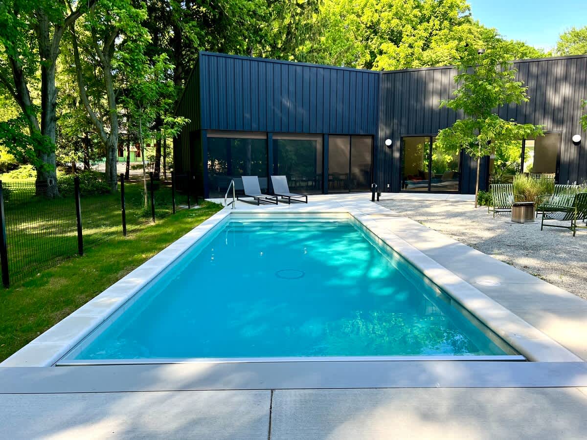 A rectangular outdoor swimming pool with clear blue water, surrounded by a concrete deck and a gravel area. The modern black house with large windows and doors forms the backdrop, set amidst lush green trees under a clear sky.