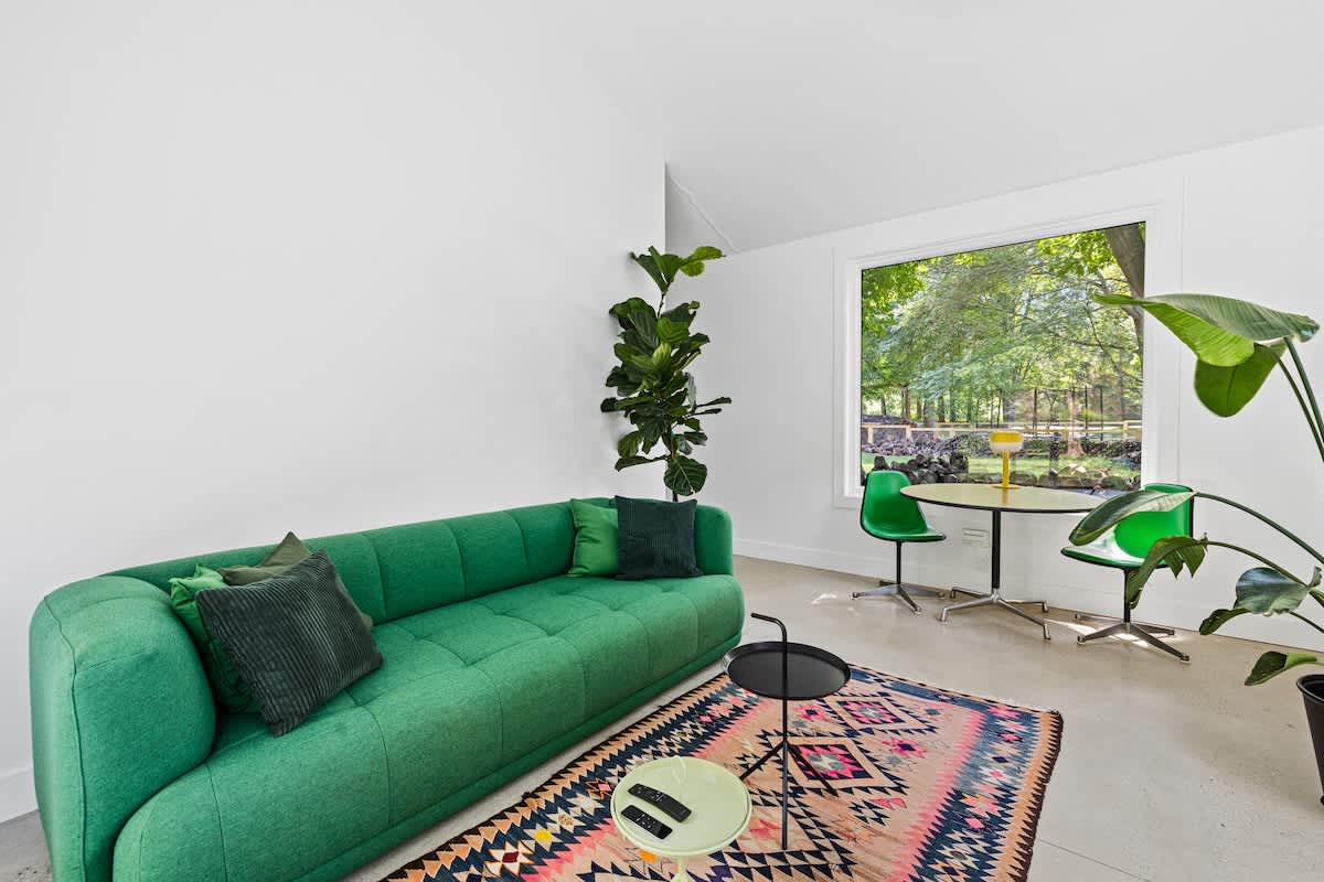 Bright living room featuring a long, tufted green sofa, a small round dining table with two green chairs, and a large window overlooking a verdant landscape. A tall fiddle leaf fig tree and a patterned rug complete the space.