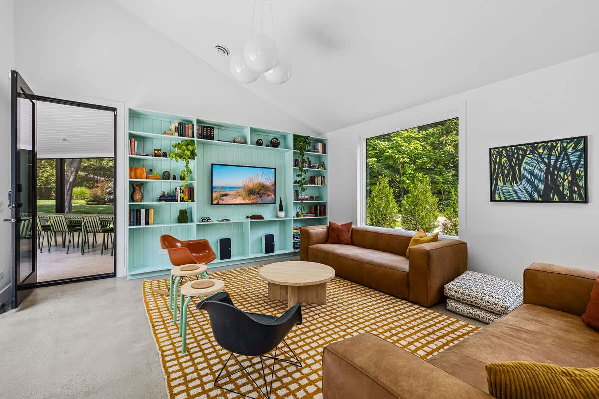 A cozy living area with a large built-in bookshelf painted in a light teal color, housing a TV and various decor items. Two brown leather sofas and a yellow patterned rug define the seating space, with large windows offering garden views.