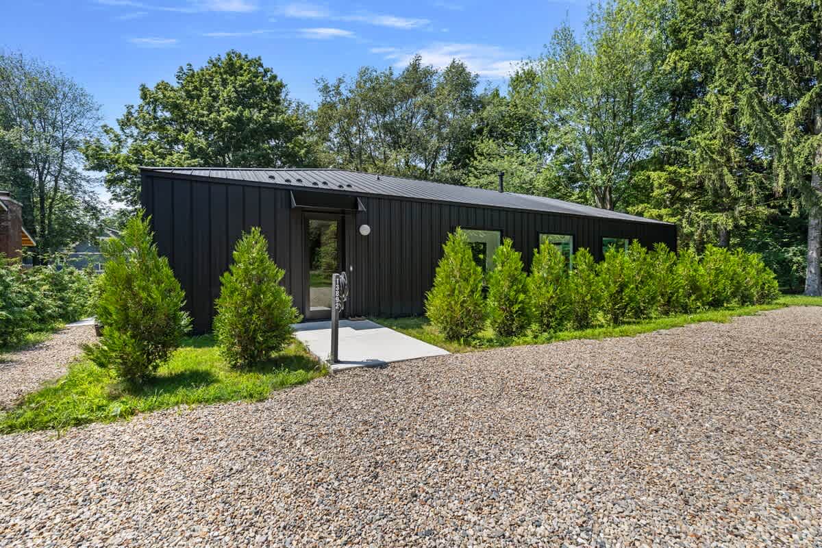 Modern black-clad house with a minimalist design, featuring a gravel driveway and small evergreen shrubs lining the front. The entrance is marked by a concrete path leading to a glass door.