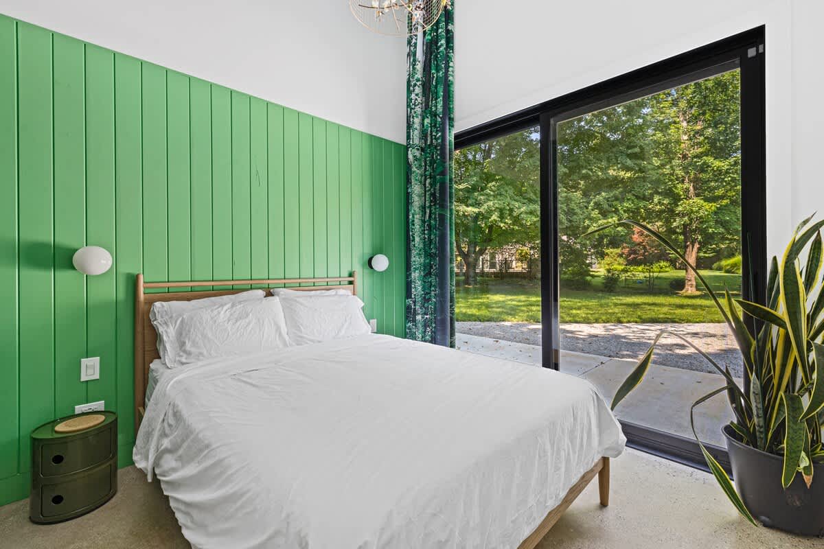 Spacious bedroom with a white bed, a vibrant green shiplap accent wall, and a large sliding glass door providing abundant natural light and views of the outdoor garden. A tall snake plant adds a natural element to the corner.