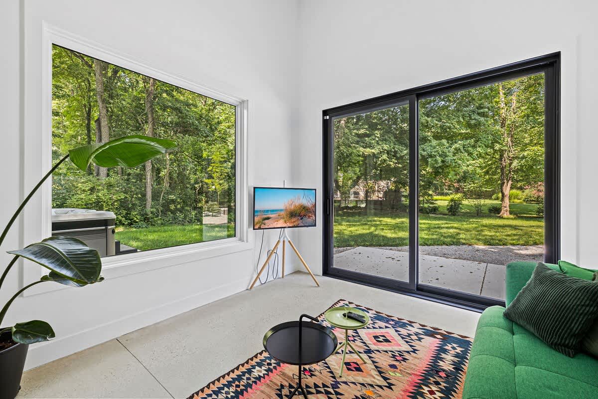 Modern living space with white walls, concrete floor, a green sofa, and large windows offering lush green outdoor views. A small TV on a tripod stand and a patterned rug add character.