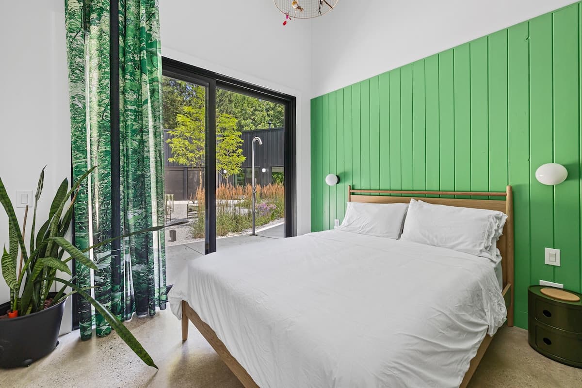 Bright bedroom featuring a light wood bed frame with white bedding, a vibrant green vertical plank accent wall, and a large sliding glass door opening to an outdoor area.