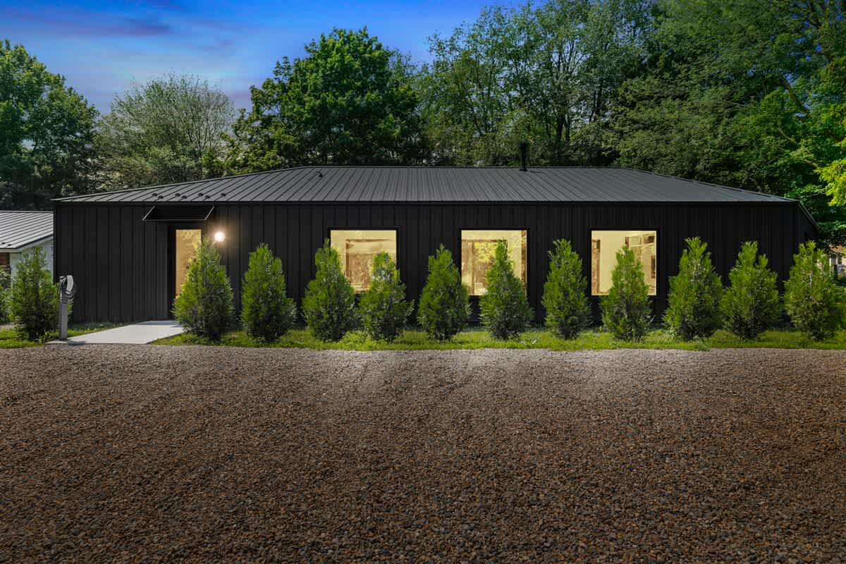 Modern, dark-clad single-story house with a low-sloping roof, featuring several square windows and a row of small evergreen trees in front of a large gravel driveway.