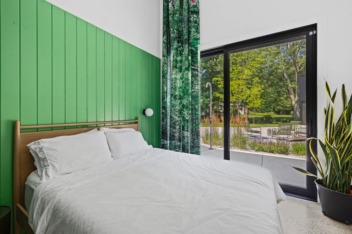 A modern bedroom with a wooden bed frame, crisp white bedding, and a vibrant green slatted accent wall. A large sliding glass door opens to an outdoor patio with lush greenery, complemented by a tall snake plant in a black pot.
