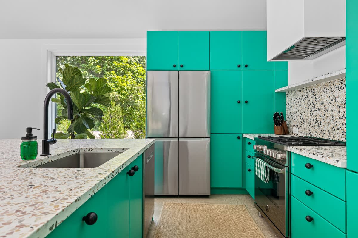 Detailed view of a modern kitchen featuring bright teal cabinets, speckled white countertops, a stainless steel refrigerator, and a gas range, with a black faucet over the sink and a large window providing natural light.