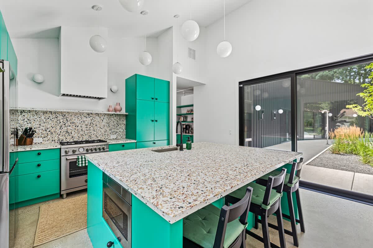 Vibrant kitchen with bright green cabinetry, speckled terrazzo countertops and backsplash, and a stainless steel range. White globe pendant lights hang from the high ceiling.
