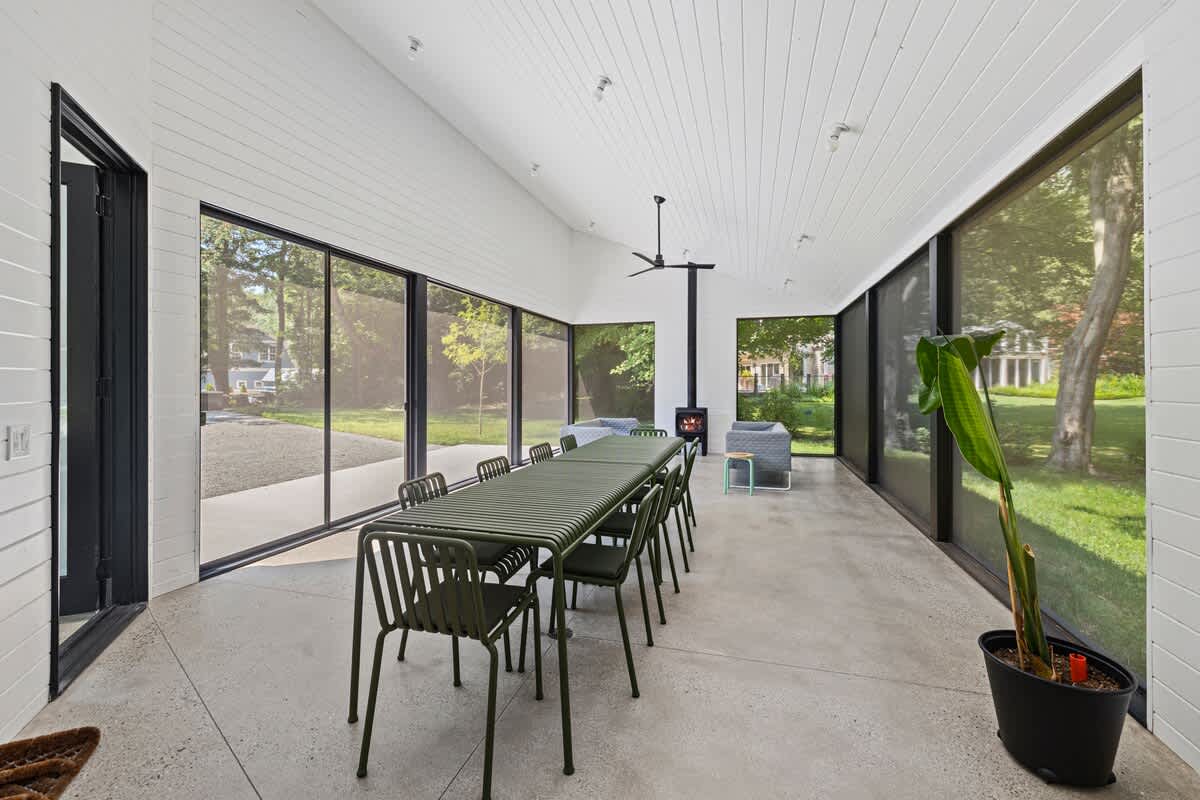 Modern dining room with a long green slatted table and matching chairs, concrete floor, and floor-to-ceiling windows offering garden views. A black wood-burning stove is visible in the background.