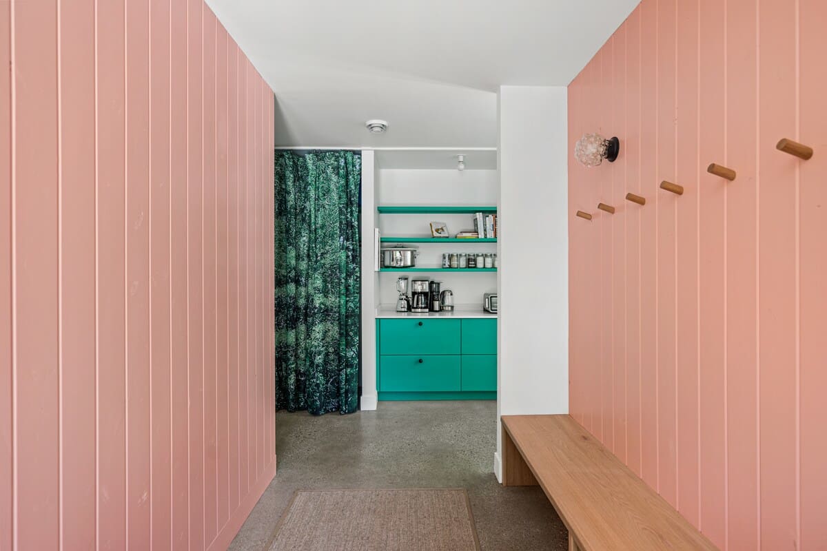 Hallway with pink vertical paneling on the walls, a wooden bench, and coat hooks, leading to a small teal kitchenette area with shelves, a coffee maker, and a green forest-patterned curtain.