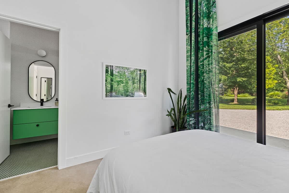 Bright bedroom featuring a white bed and a large sliding glass door offering views of the lush outdoor garden. A modern bathroom with a green vanity is visible through the open doorway, complementing the room's fresh aesthetic.