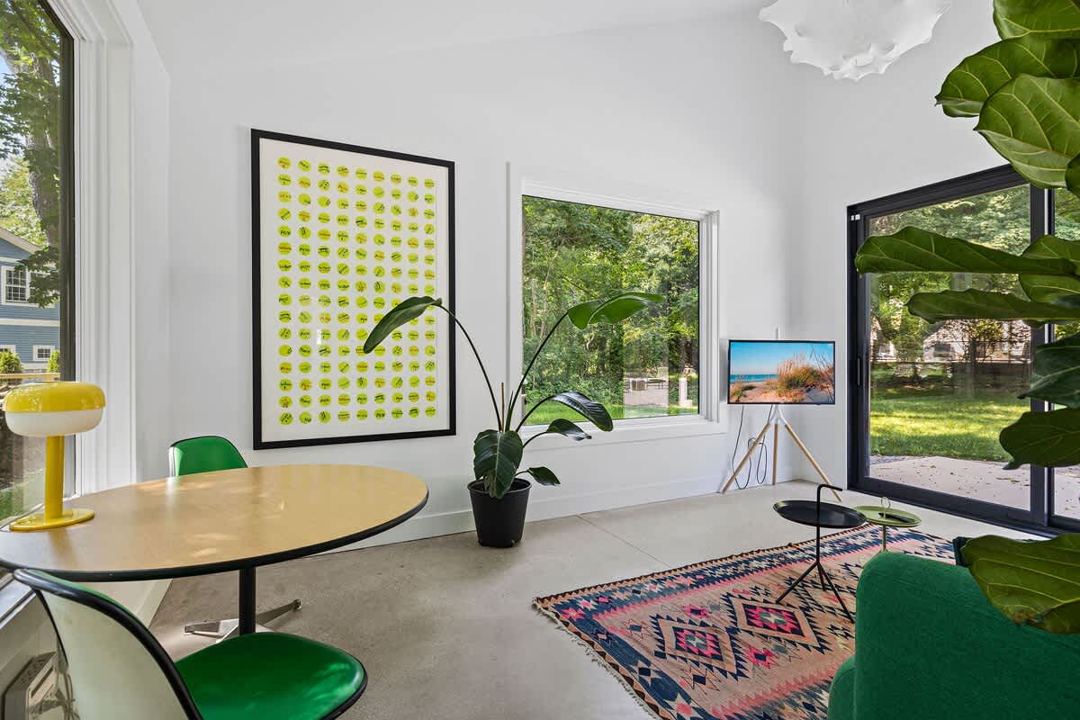A bright sitting area with a light wood oval table and green chairs, positioned next to a large window overlooking lush greenery. A vibrant yellow smiley face art piece hangs on the wall, and a modern TV on a tripod stand is visible.