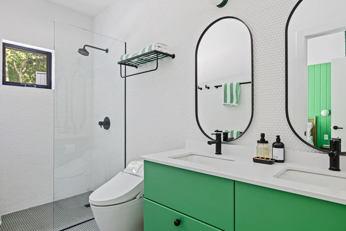 Contemporary bathroom featuring a double vanity with bright green cabinets, white countertops, and black oval mirrors. The walls are adorned with white penny tiles, and a spacious walk-in shower with a black-framed window is visible.